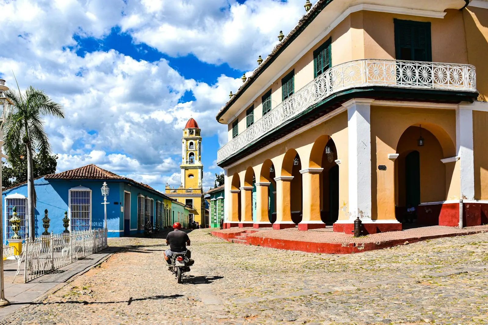 Photo by AXP Photography A motorcyclist rides down sunlit cobblestone streets in a colorful Cuban town, showcasing colonial architecture.